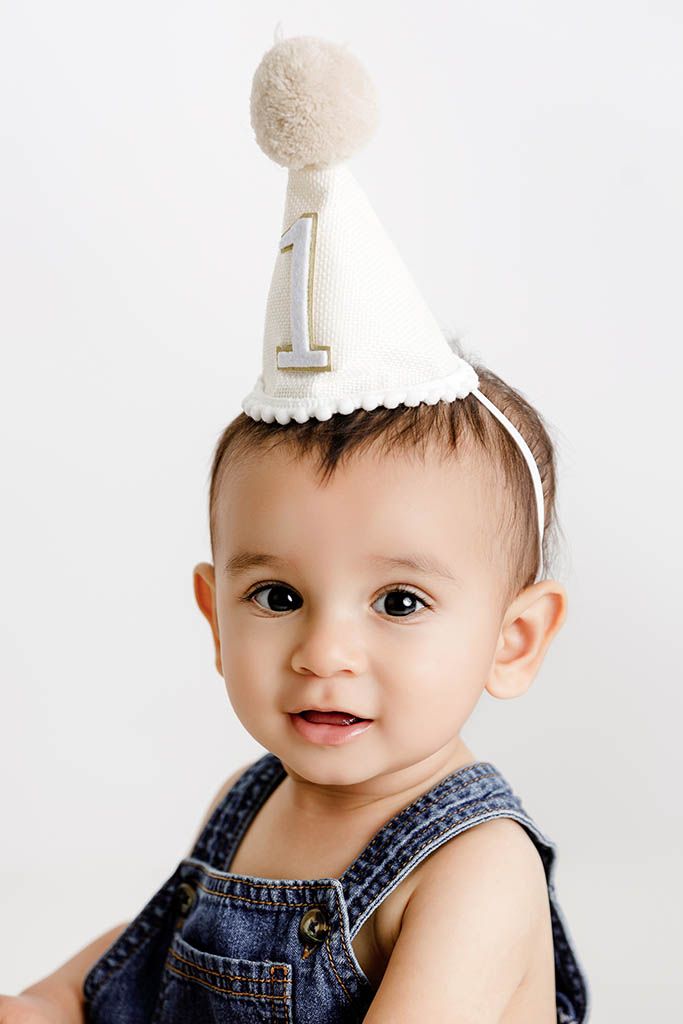 Adorable baby wearing a white number one birthday party hat and denim overalls, celebrating their first birthday.