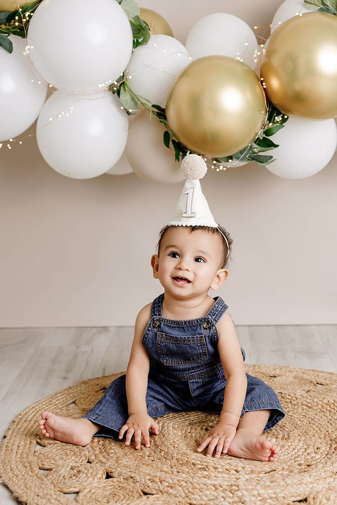 Smiling baby in denim overalls wearing a number one birthday hat sits on a woven rug with gold and white balloons.