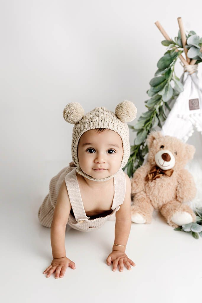 Cute baby in knit bear bonnet hat crawls on white floor near teddy bear and teepee prop in studio photo.