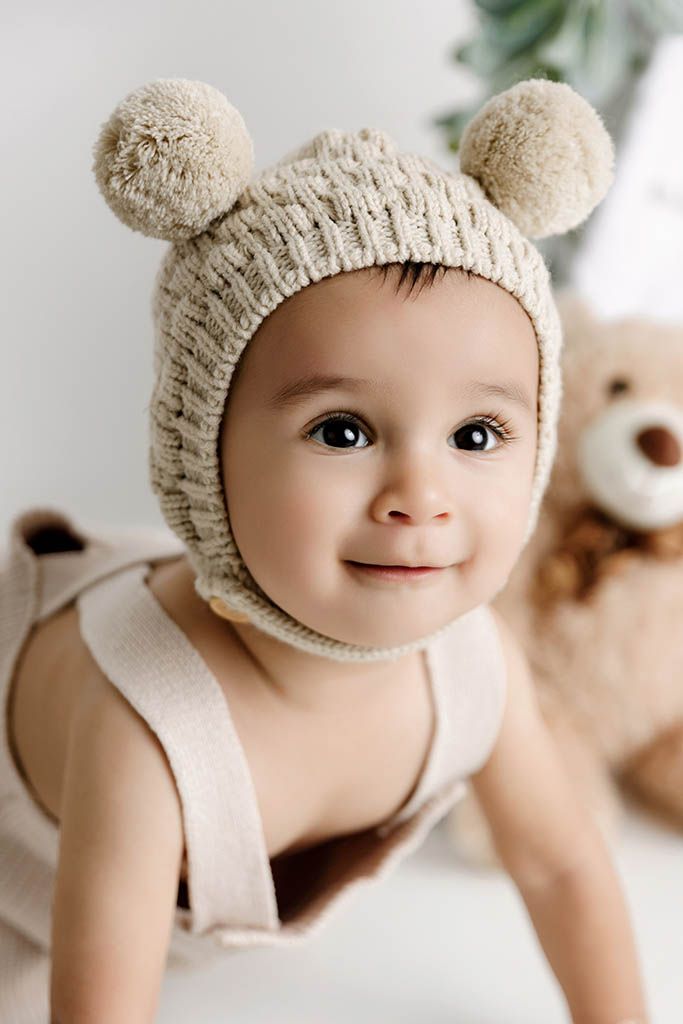 Affordable photographer Menifee photo of adorable baby wearing a beige knitted bear bonnet hat with pom pom ears, smiling cutely at camera.