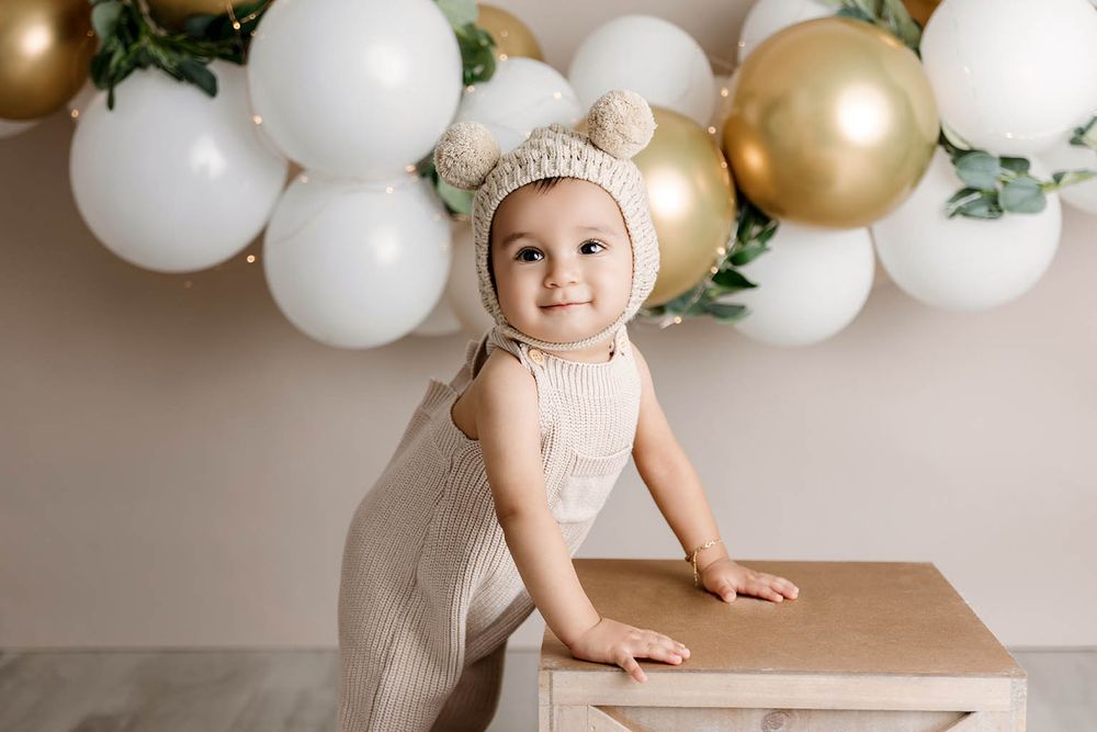 Adorable baby in bear bonnet stands at wooden table with gold and white balloon backdrop.