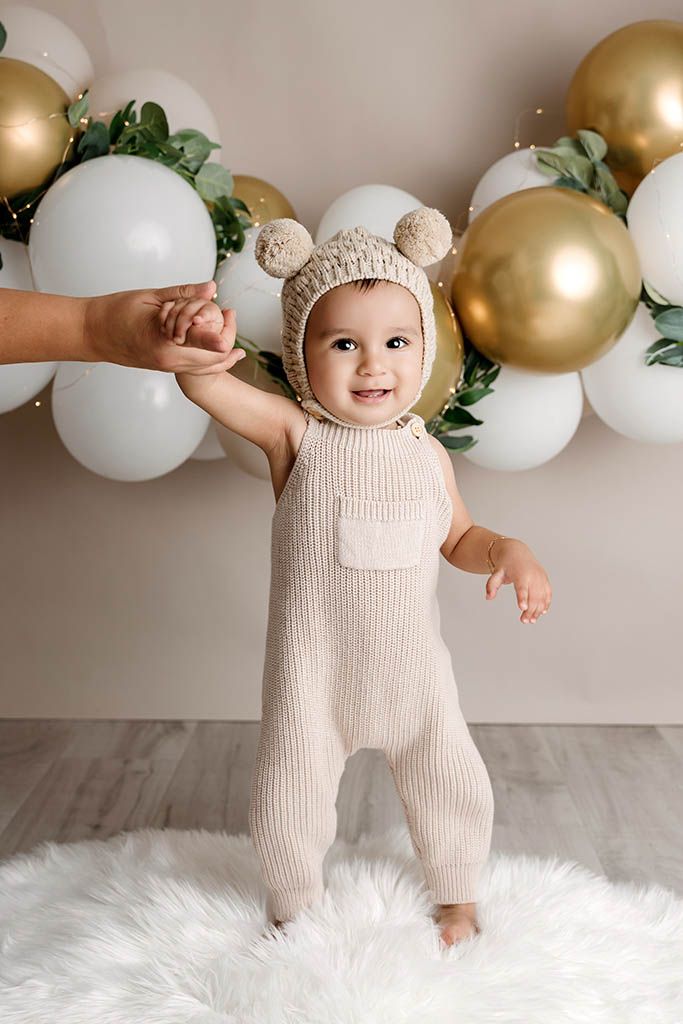 Happy baby in beige knit romper and bear bonnet standing with balloon backdrop at milestone photo shoot.