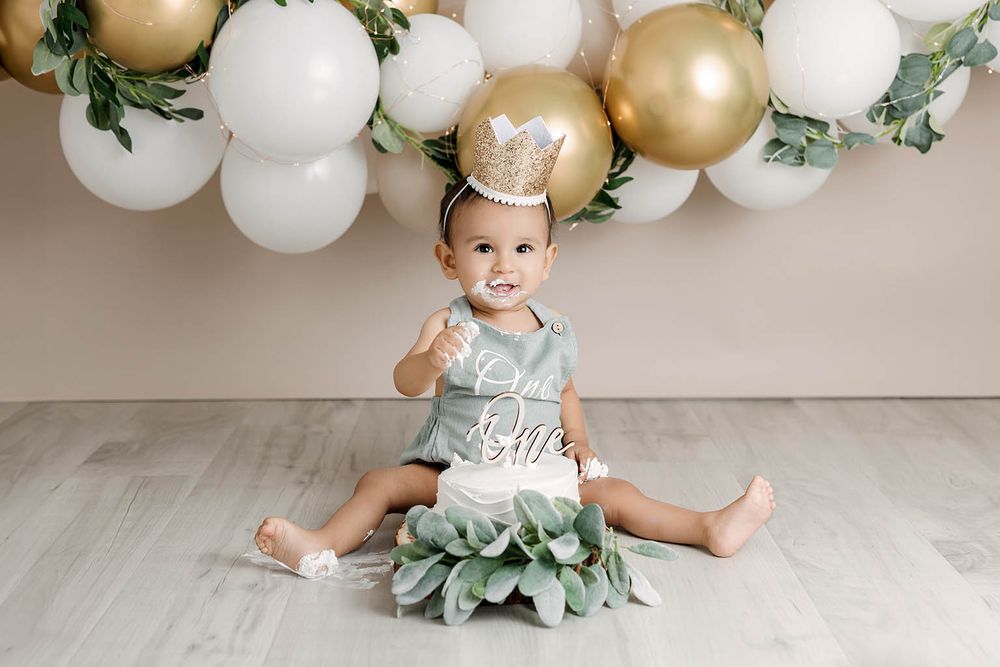 Baby in sage romper sits with a crown amid balloons and greenery for a first birthday cake smash.