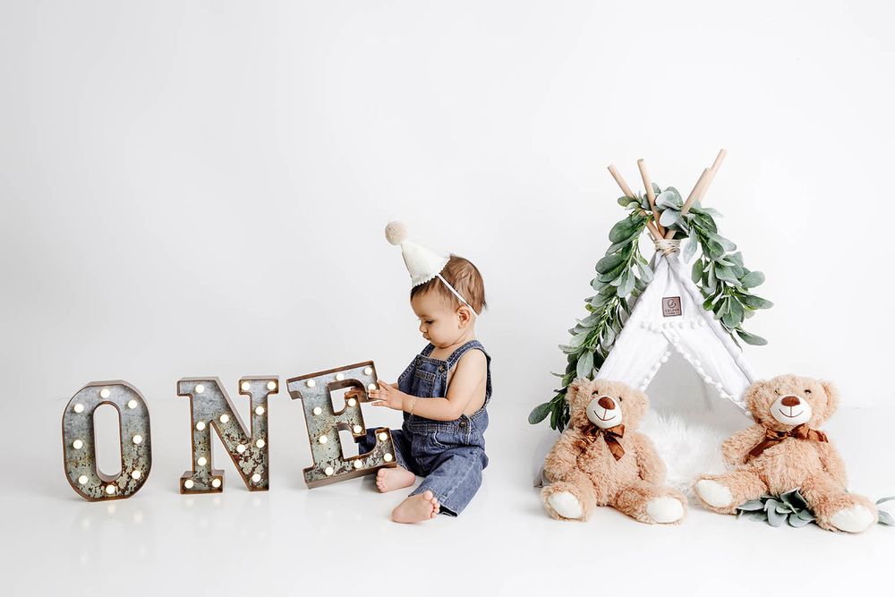Baby in denim overalls sits beside illuminated ONE letters, a small teepee, and teddy bears for a first birthday photo.