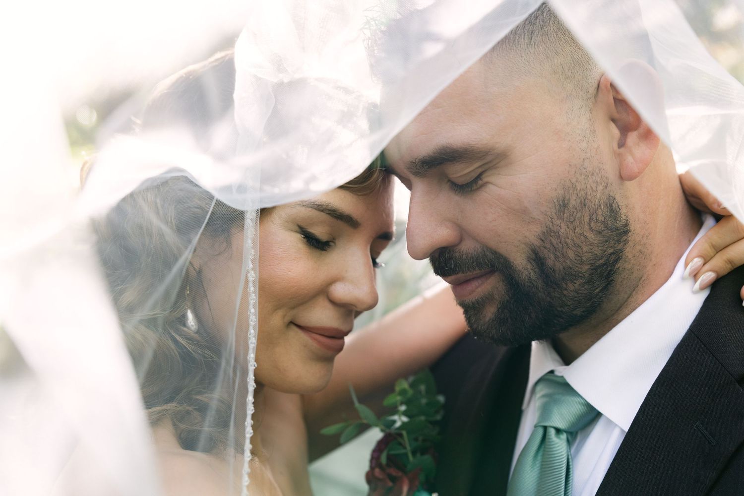 Bride and groom share an intimate moment under a wedding veil, foreheads touching, in a romantic outdoor portrait.