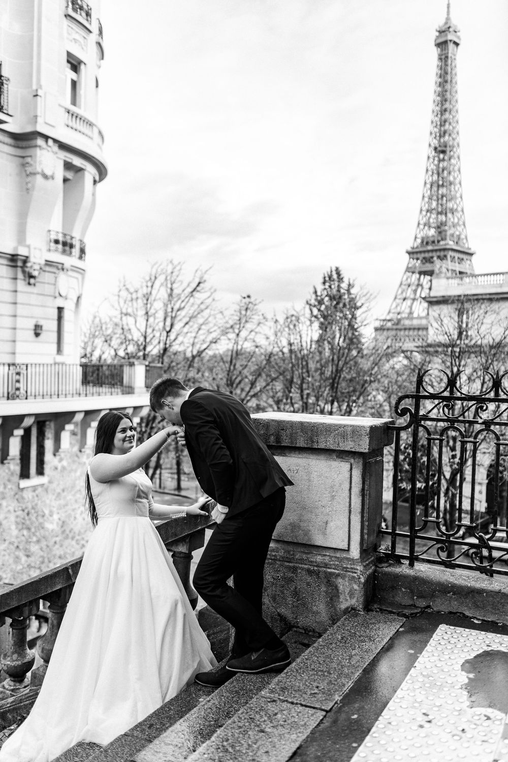 Black and white photo of bride and groom on Paris balcony with Eiffel Tower in background.