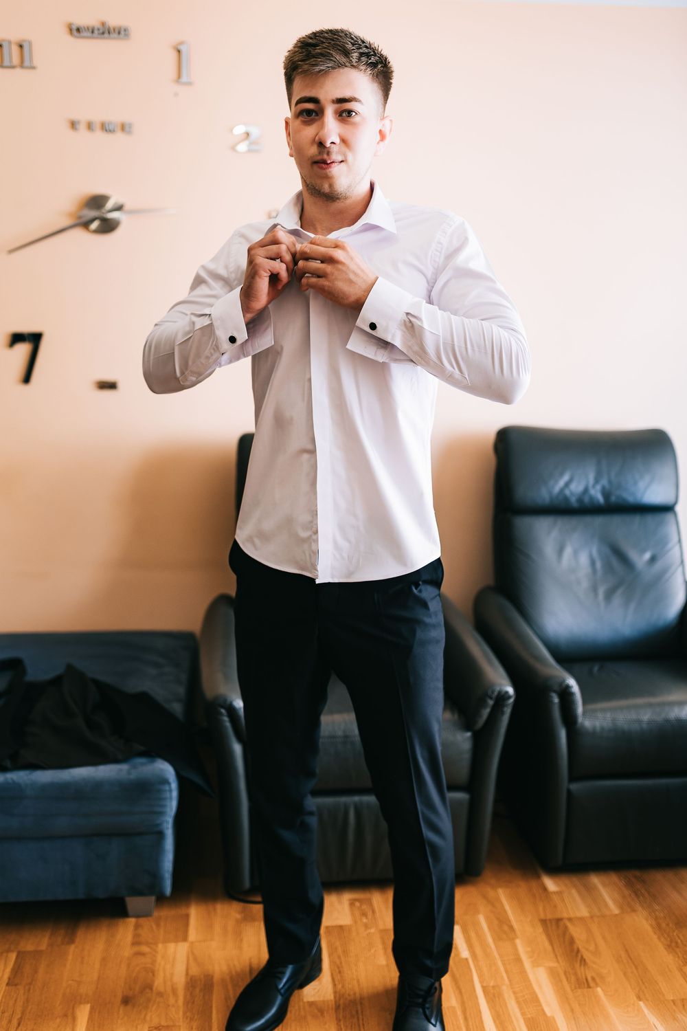 Handsome groom in white dress shirt and black trousers buttoning up while getting ready for wedding.