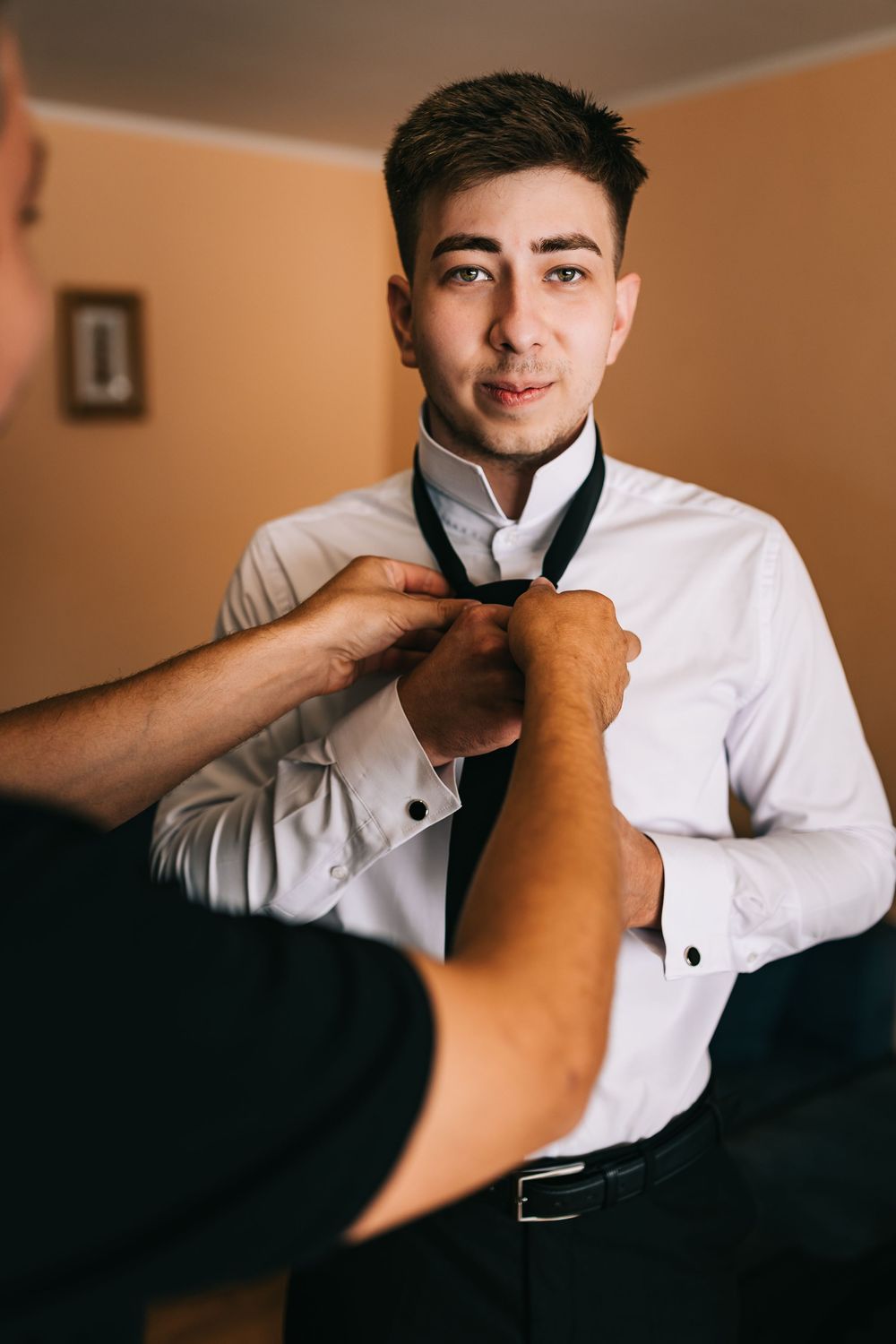Groomsman helping groom adjust black tie while getting ready for wedding ceremony indoors.