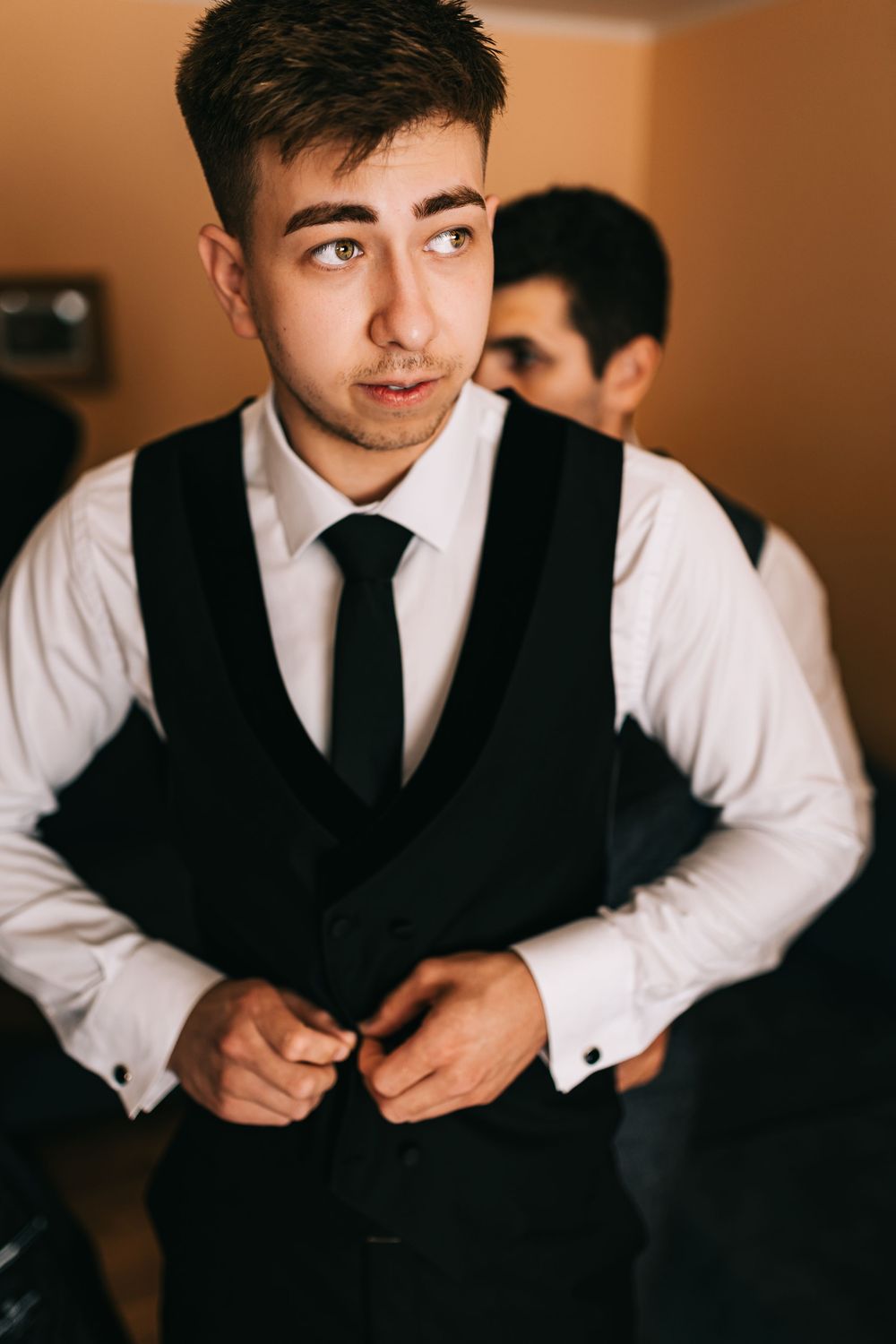 Young groom buttoning his black vest while getting ready for his wedding ceremony indoors.