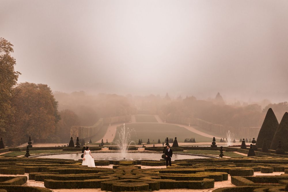 Bride and groom stand in misty formal gardens of Palace of Versailles with fountain.