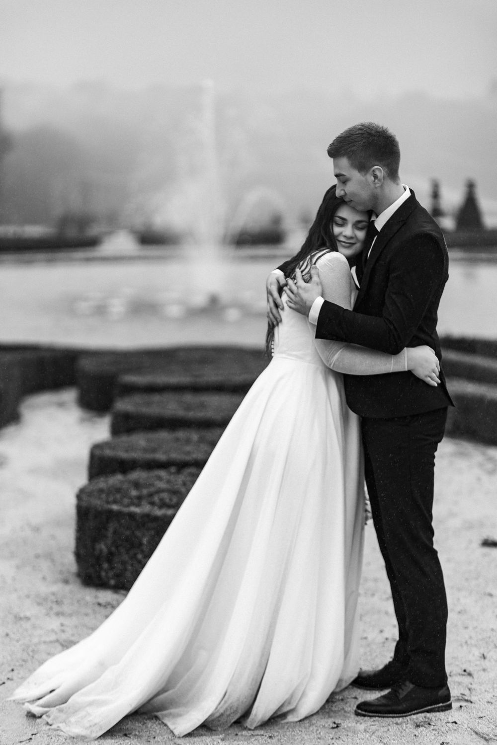 Groom tenderly embraces bride in black and white photo at Versailles garden fountain.