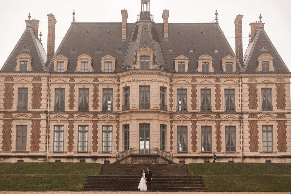 Bride and groom embrace before a grand French château with ornate brick façade and steep mansard roof.