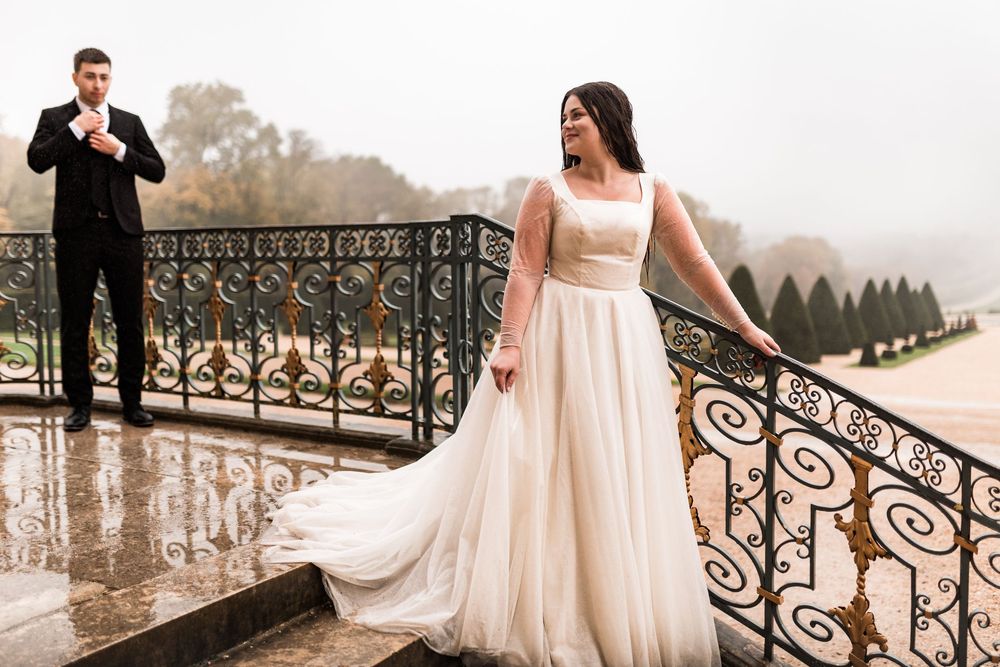 Bride in elegant white gown stands on rain-soaked balcony with groom adjusting his suit behind her.