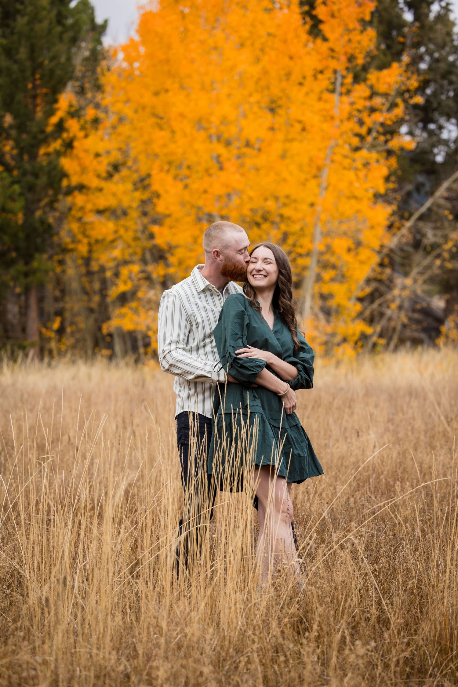 Couple embracing in golden autumn field with vibrant yellow aspen trees, woman in green dress smiling during fall engagement photo.