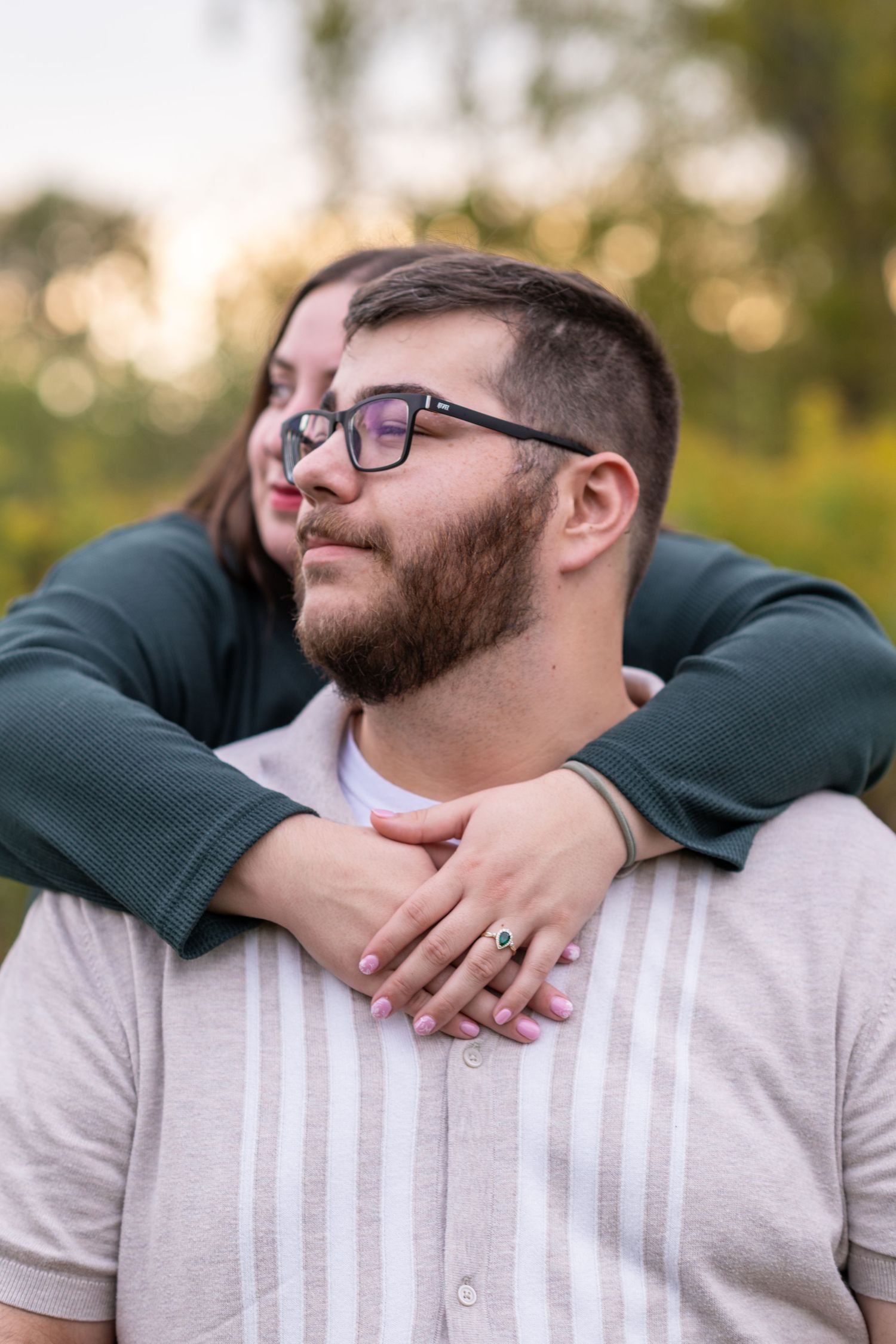 A couple embraces outdoors, woman hugging man from behind, both wearing glasses, with a ring visible on her hand.