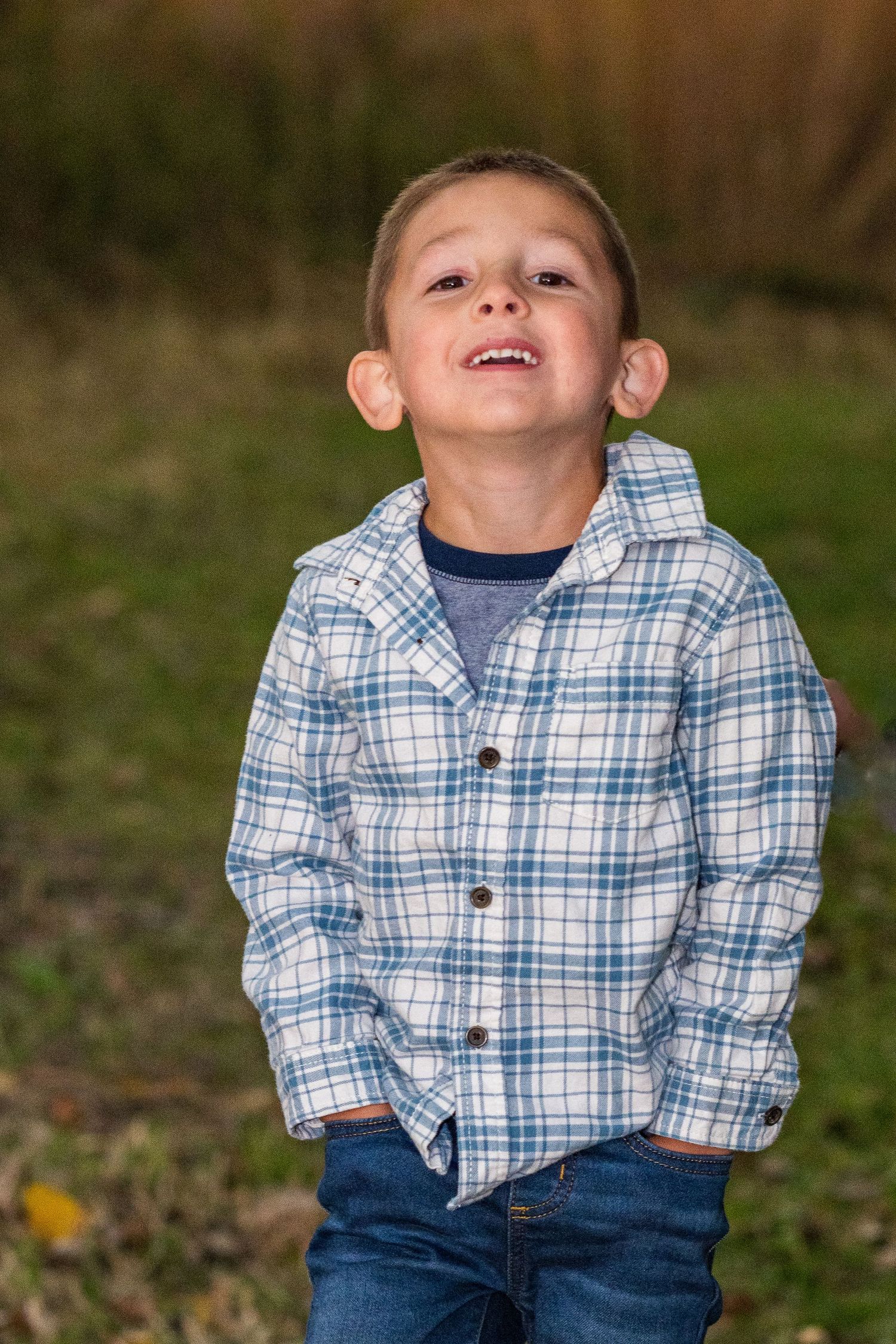 Young boy in plaid shirt and jeans smiling outdoors with hands in pockets against green grass background.