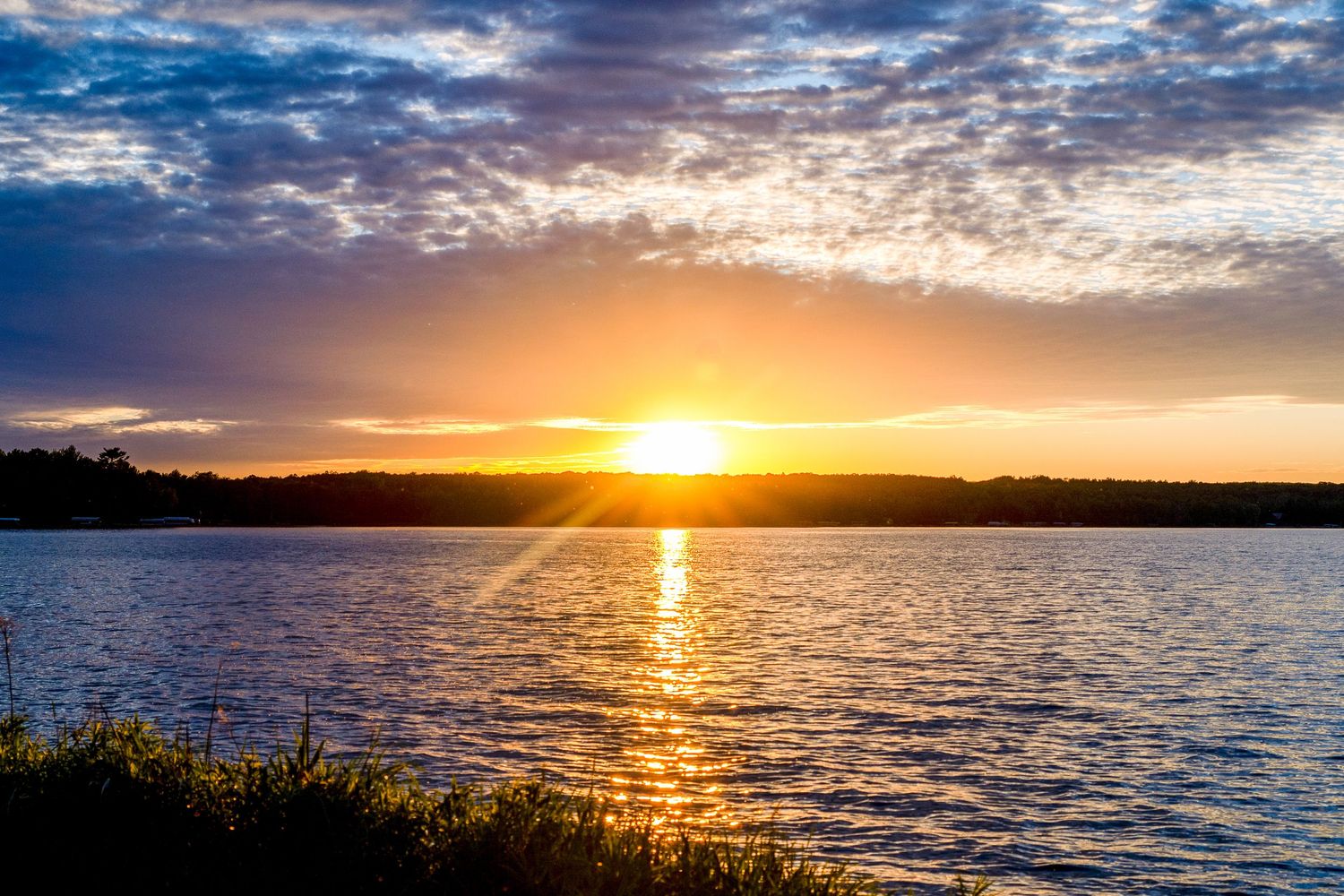 Golden sunset over a calm lake with vibrant orange sky, reflected sunlight on water, and silhouetted trees on the horizon.