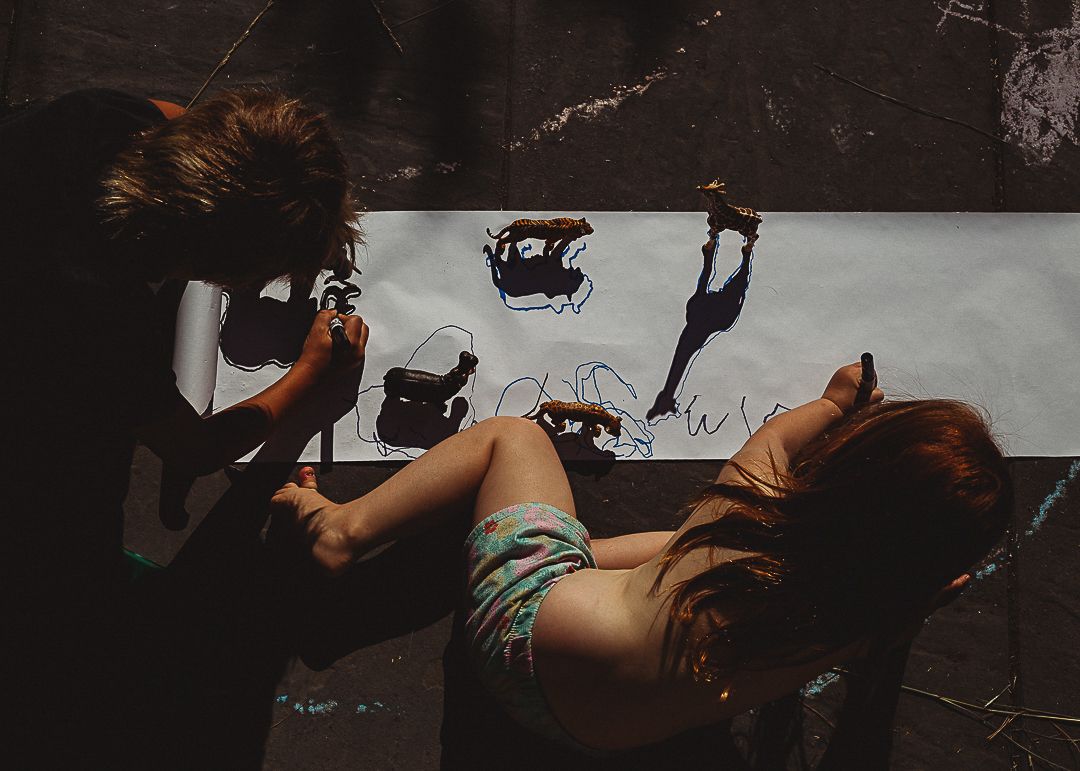 Two children drawing monster illustrations on large white paper spread on the floor.