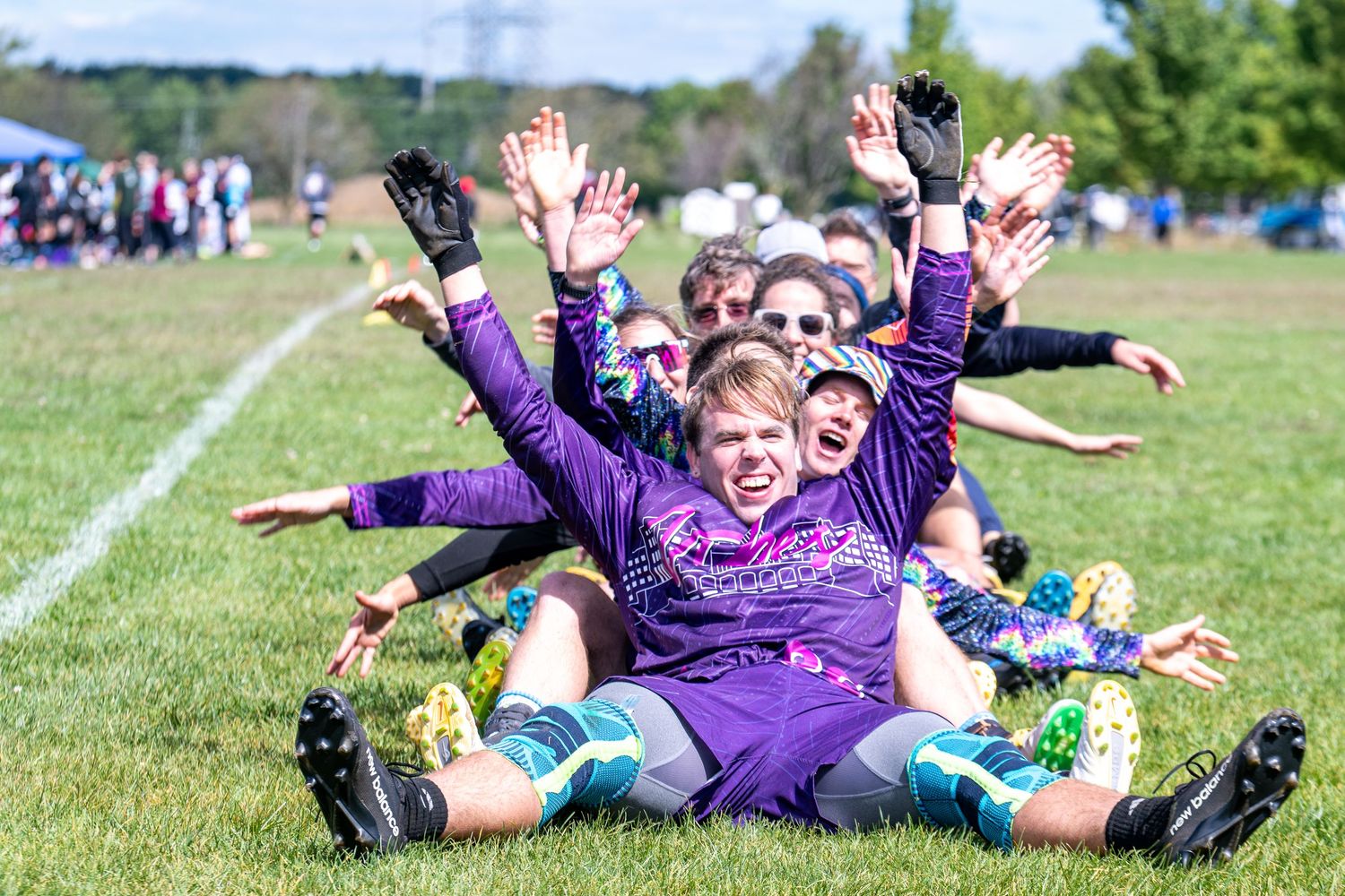 Women's soccer team celebrating on grass field, wearing purple uniforms, sitting together with hands raised joyfully.