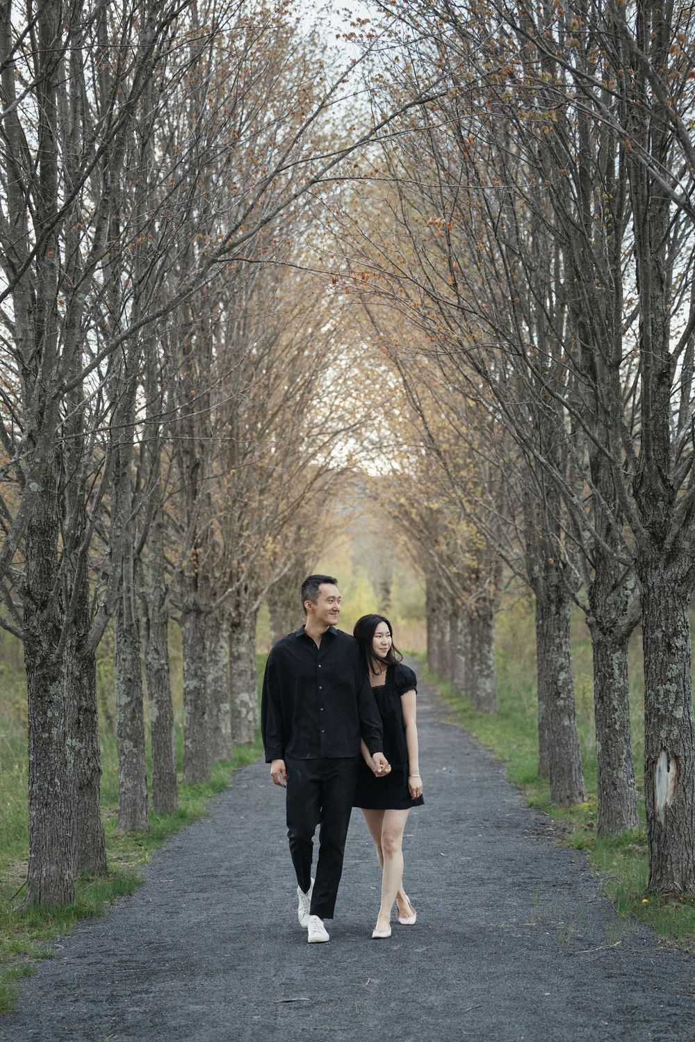 Couple in matching black outfits walking hand in hand along a tree-lined path in autumn.