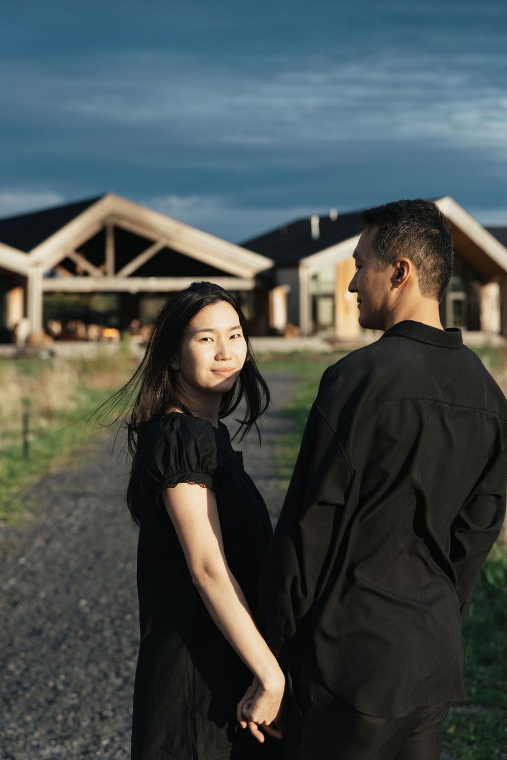 A couple in black outfits hold hands outdoors near modern buildings under a dramatic cloudy sky at dusk.