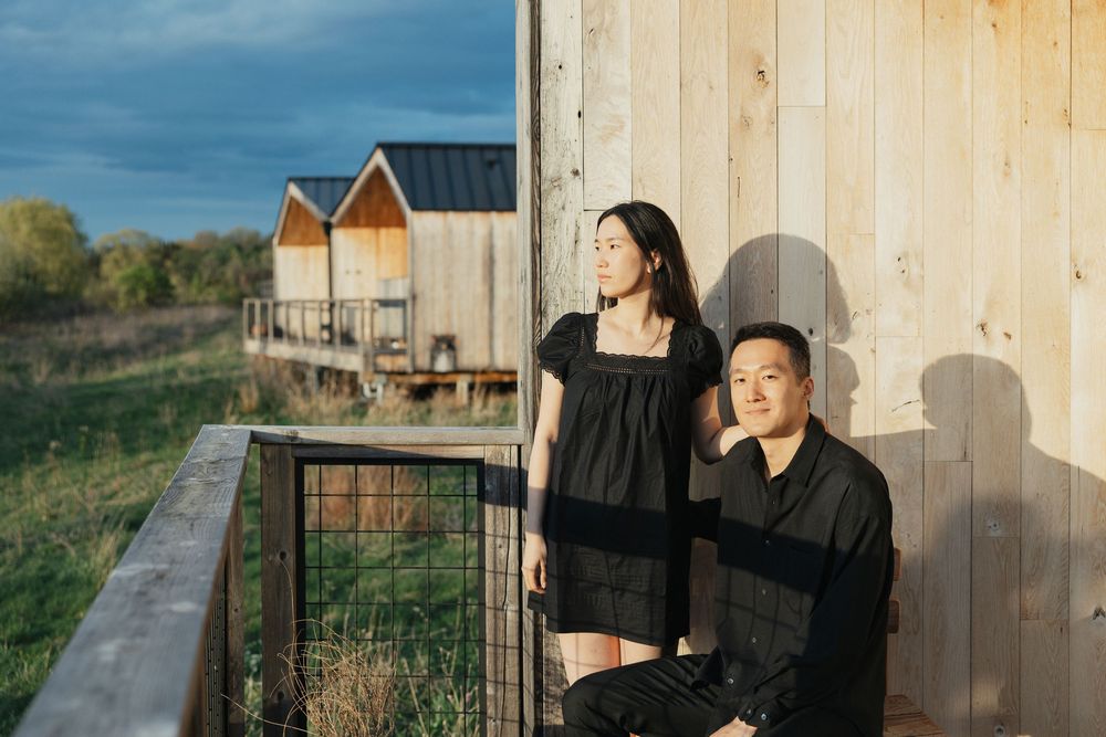 A couple in black outfits poses near a wooden barn building surrounded by green fields under a dramatic cloudy sky.
