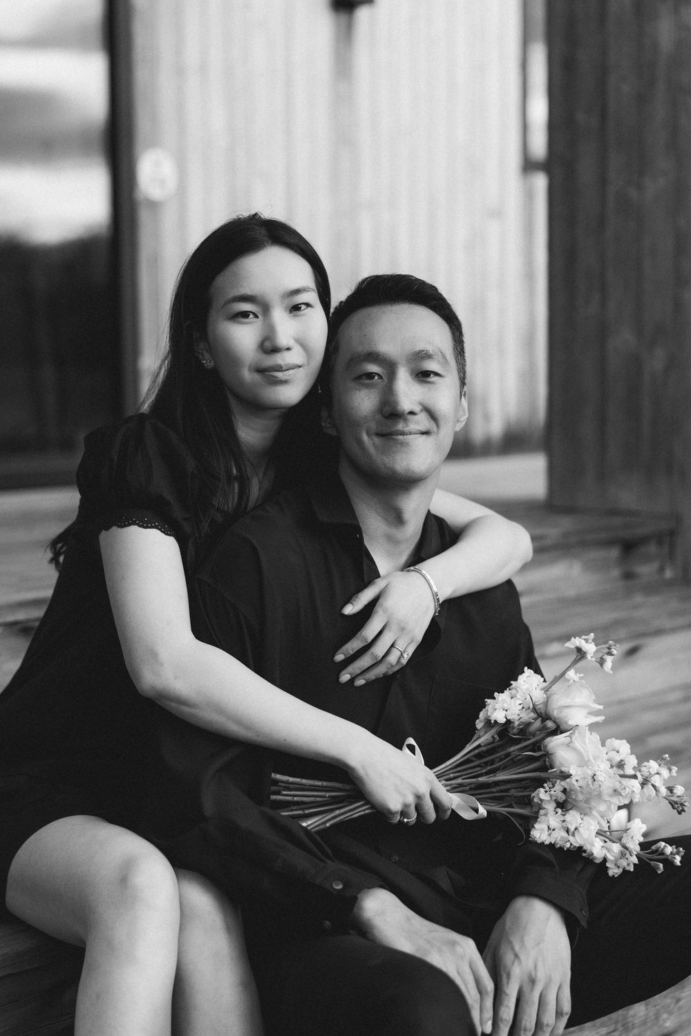 Black and white photo of a couple sitting together, woman embracing man holding a bouquet of flowers.