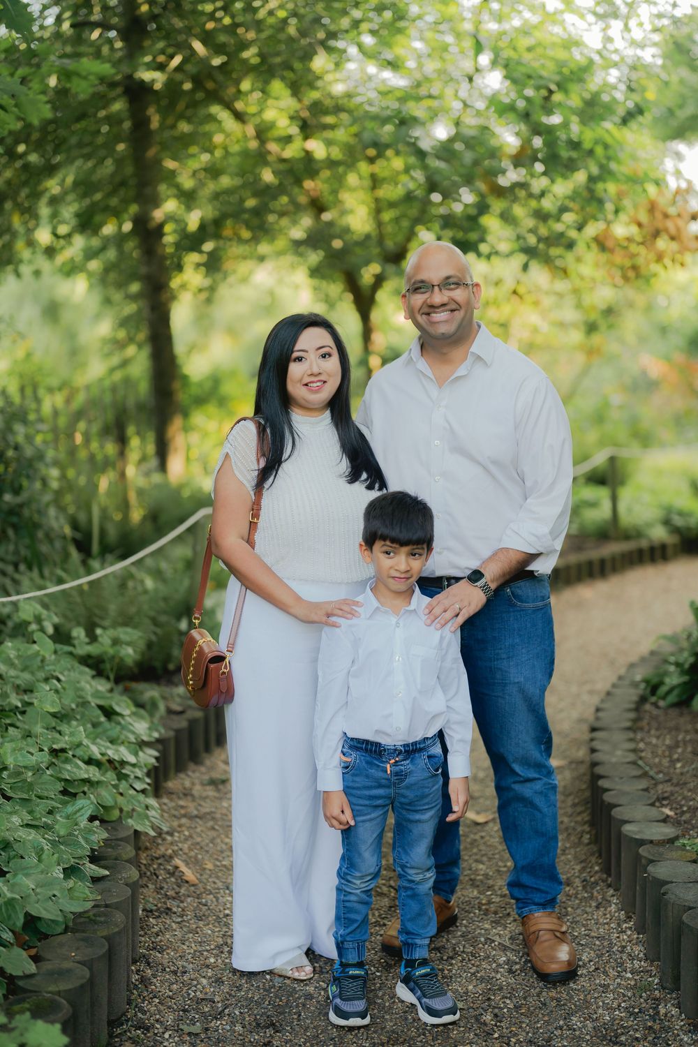 Family of three posing on a garden path, parents in white outfits with young son, surrounded by lush green trees.