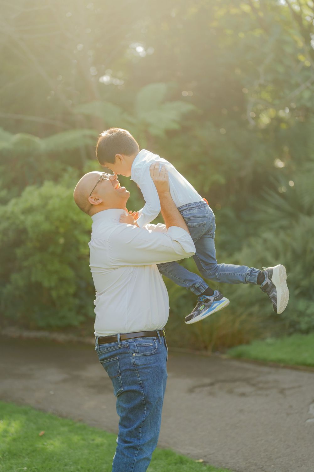 A bald grandfather in glasses lifts a young boy joyfully in a sunny green park, both smiling and sharing a playful moment.