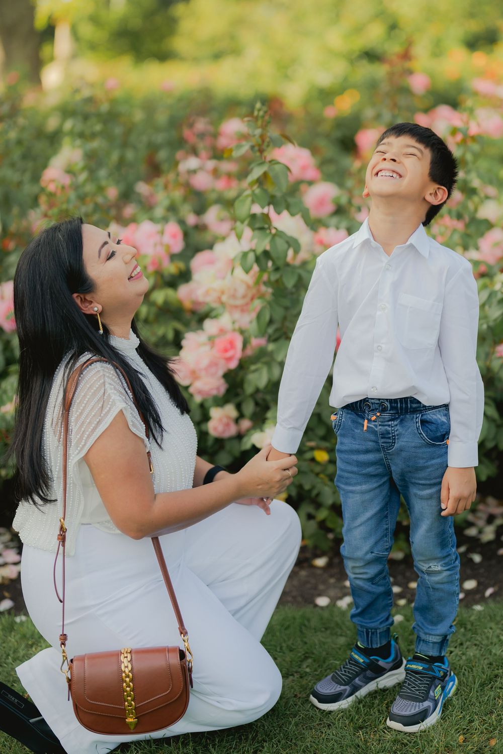 Mother kneeling beside laughing young boy in front of blooming pink roses in a sunny garden.
