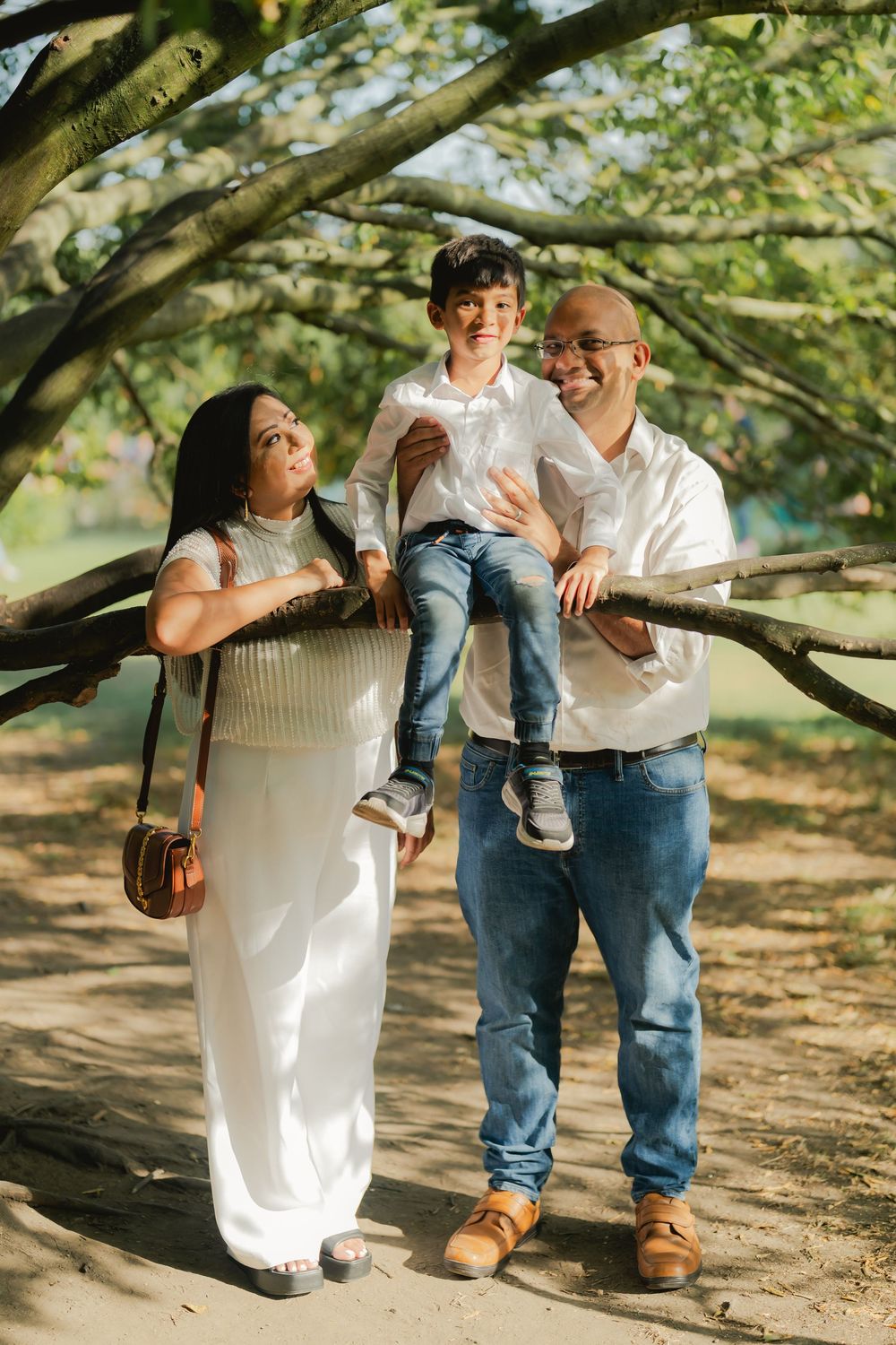 A happy South Asian family with a young boy sitting on a wooden fence rail outdoors in a sunlit park.