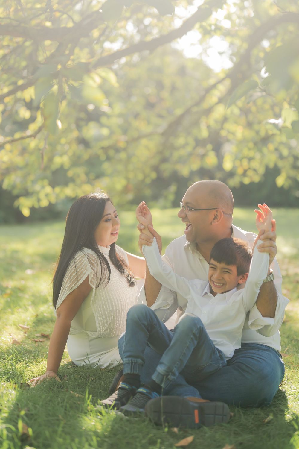 Happy family sitting on grass in sunny park, parents holding son's raised hands while smiling together outdoors.