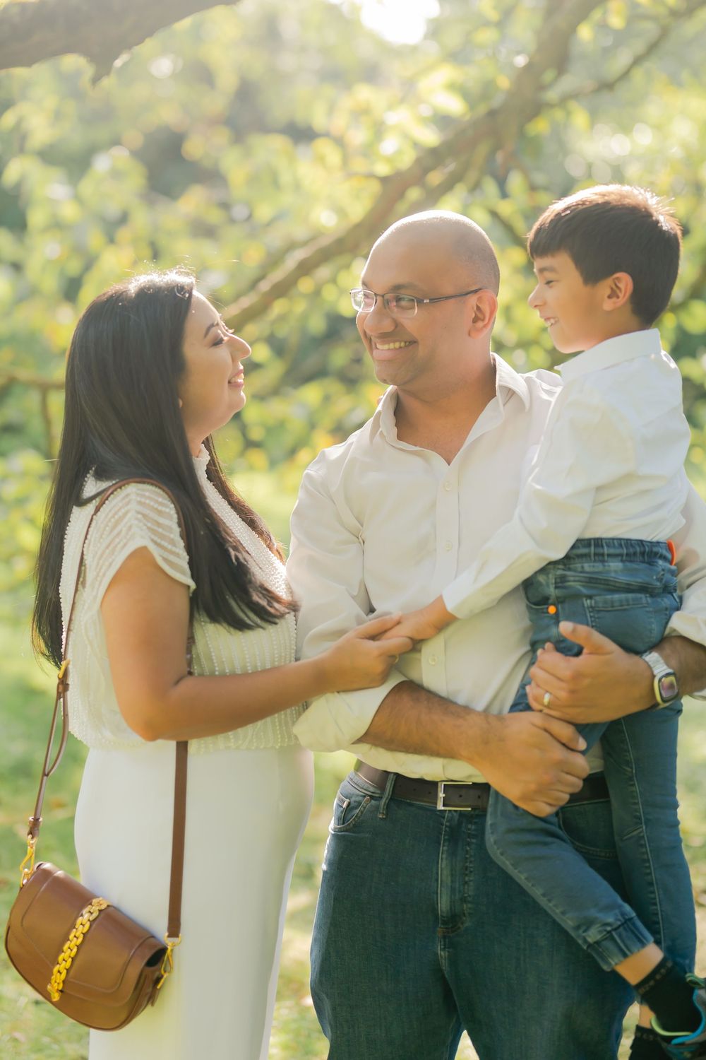 A happy family of three enjoying a sunny day outdoors, with parents smiling at their young son in a park.
