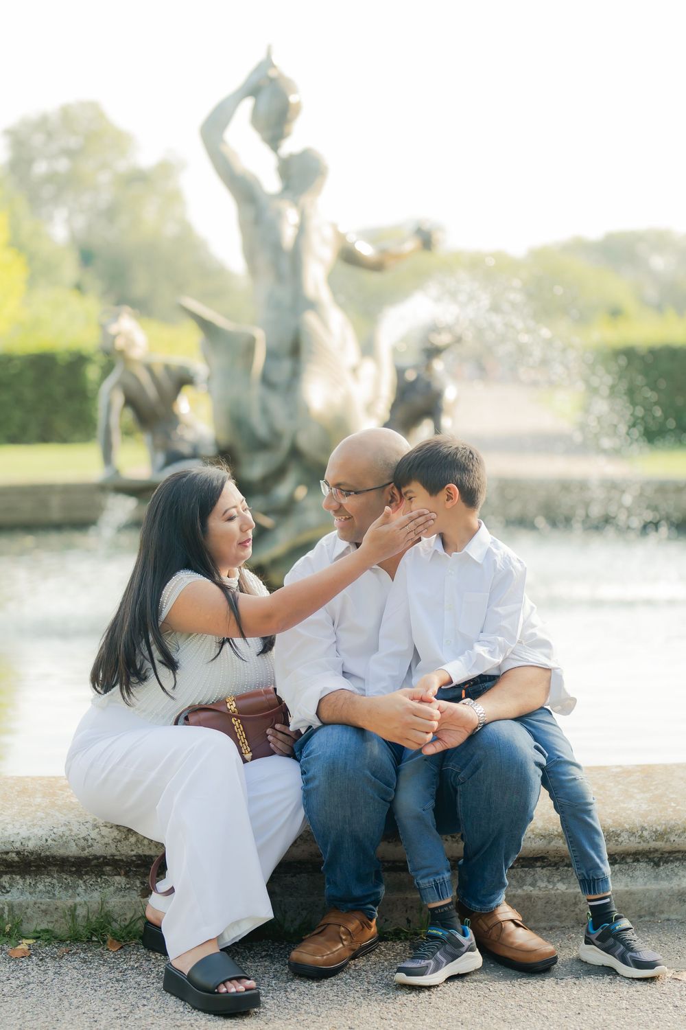Family of three sitting by an ornate fountain sculpture, sharing a tender moment in a sunlit garden setting.
