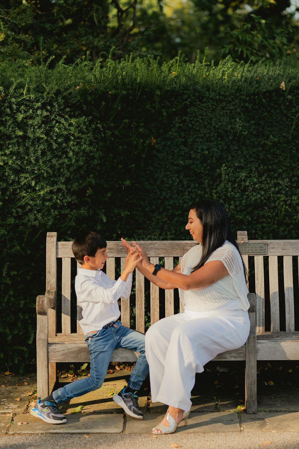 A mother and young son play a hand-clapping game on a wooden park bench, surrounded by lush green hedges.