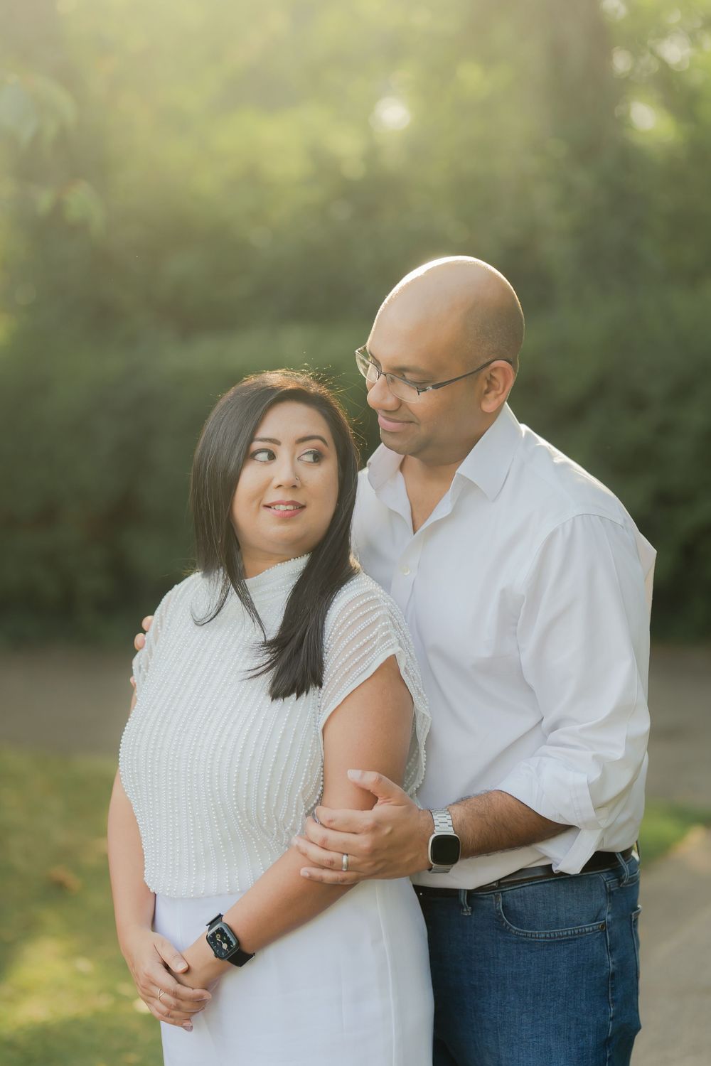 A couple in white outfits share a tender moment outdoors, with soft golden sunlight filtering through green trees behind them.
