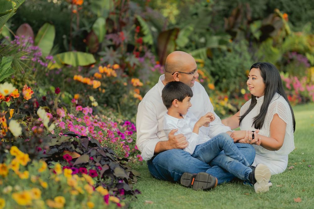Family of three sitting on grass surrounded by colorful garden flowers, sharing a warm moment together outdoors.