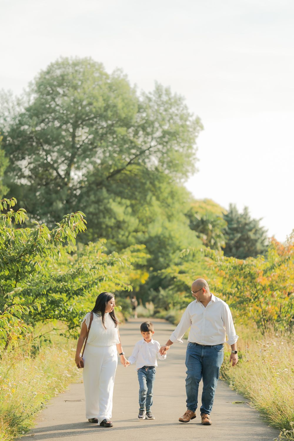 Family of three walking hand-in-hand on a scenic path surrounded by lush green trees on a sunny day.