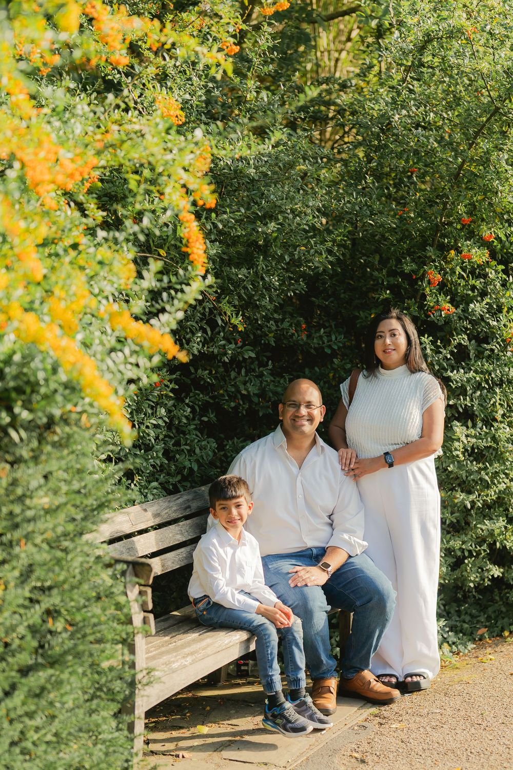Family of three posing on a wooden bench surrounded by lush green shrubs and yellow flowers in a park.