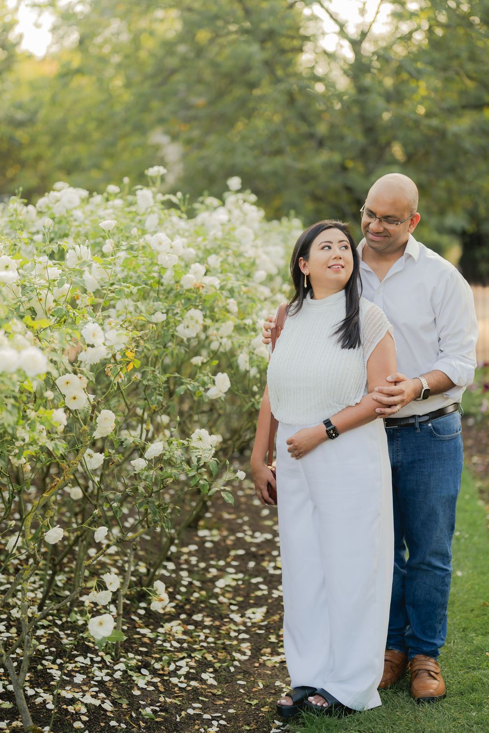 A couple embraces near white flowering bushes in a lush garden, woman in white dress, man in blue jeans and white shirt.