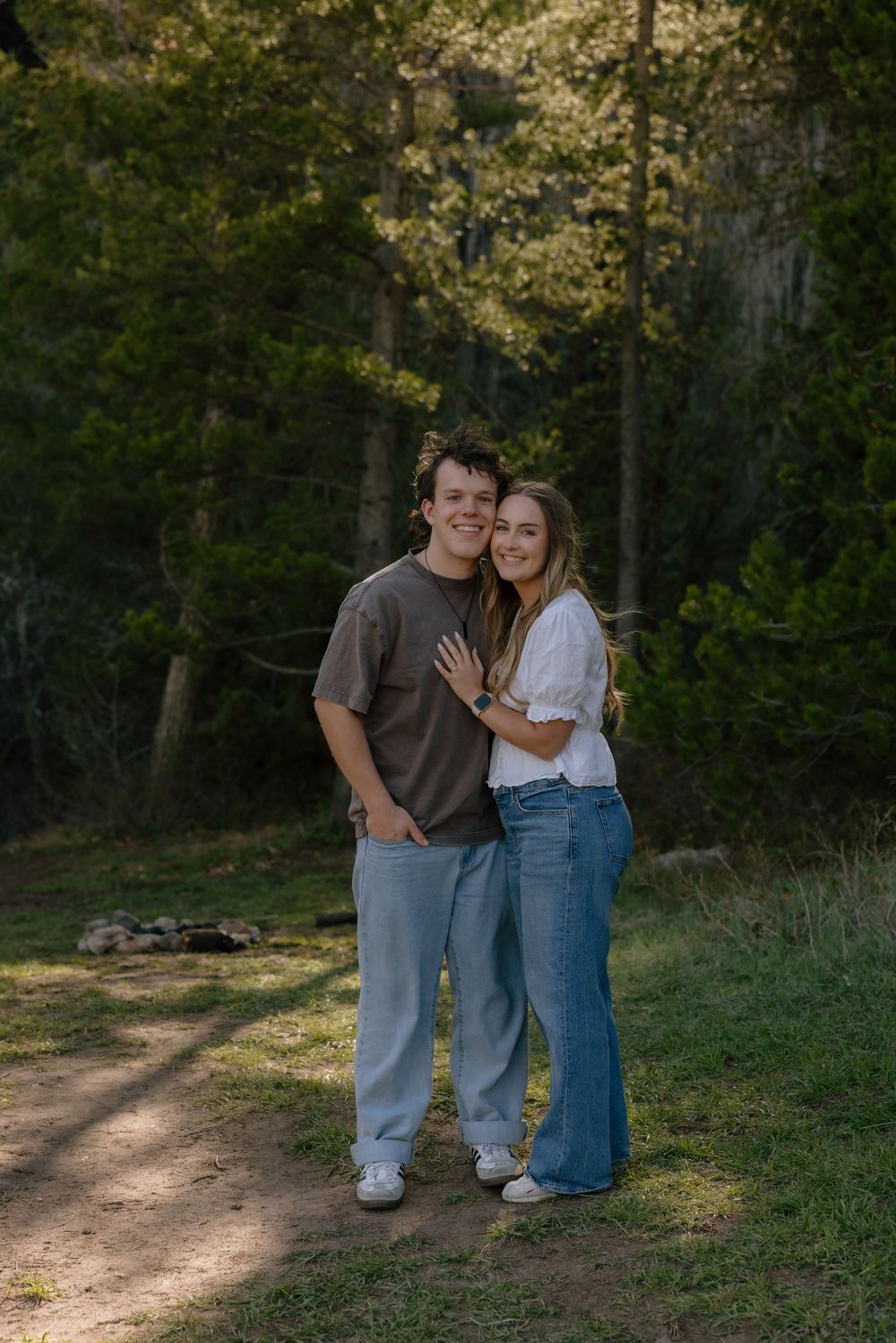 Smiling couple embracing on a sunlit forest path, woman showing engagement ring in casual outdoor setting.