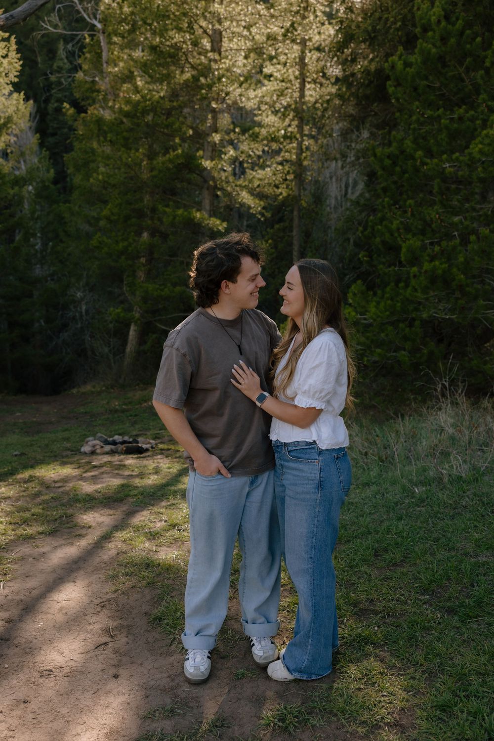 Couple smiling at each other while embracing on a forest path surrounded by tall green trees.