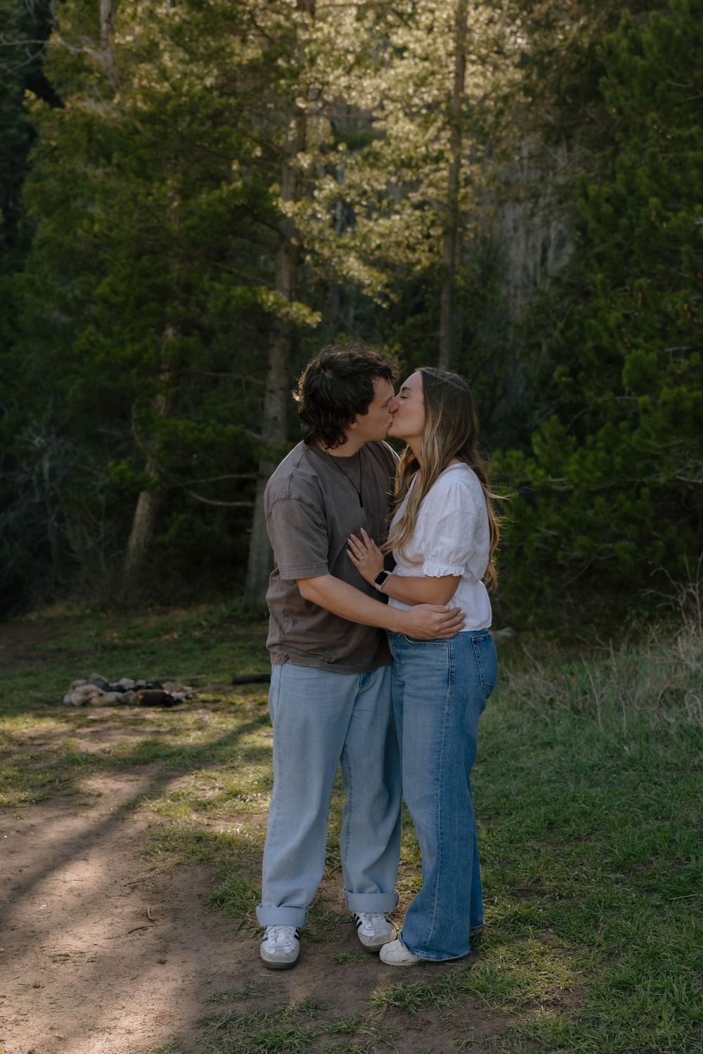 Couple sharing a romantic kiss while embracing on a forest trail surrounded by tall green trees.