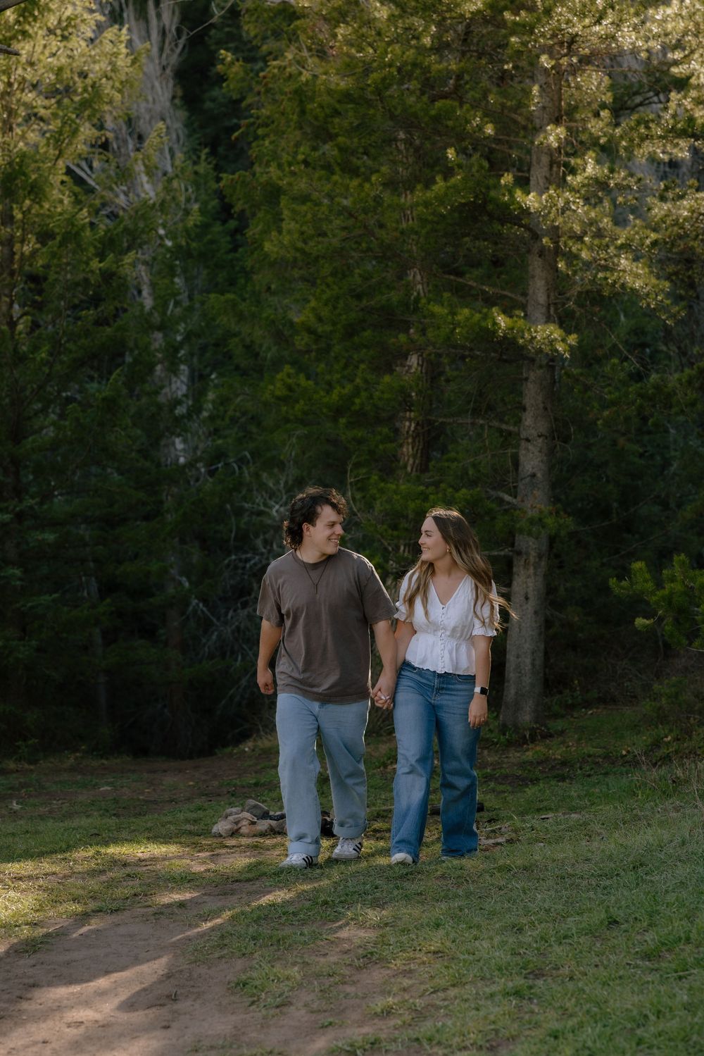 A couple holds hands while walking along a forest path surrounded by tall green trees in natural light.