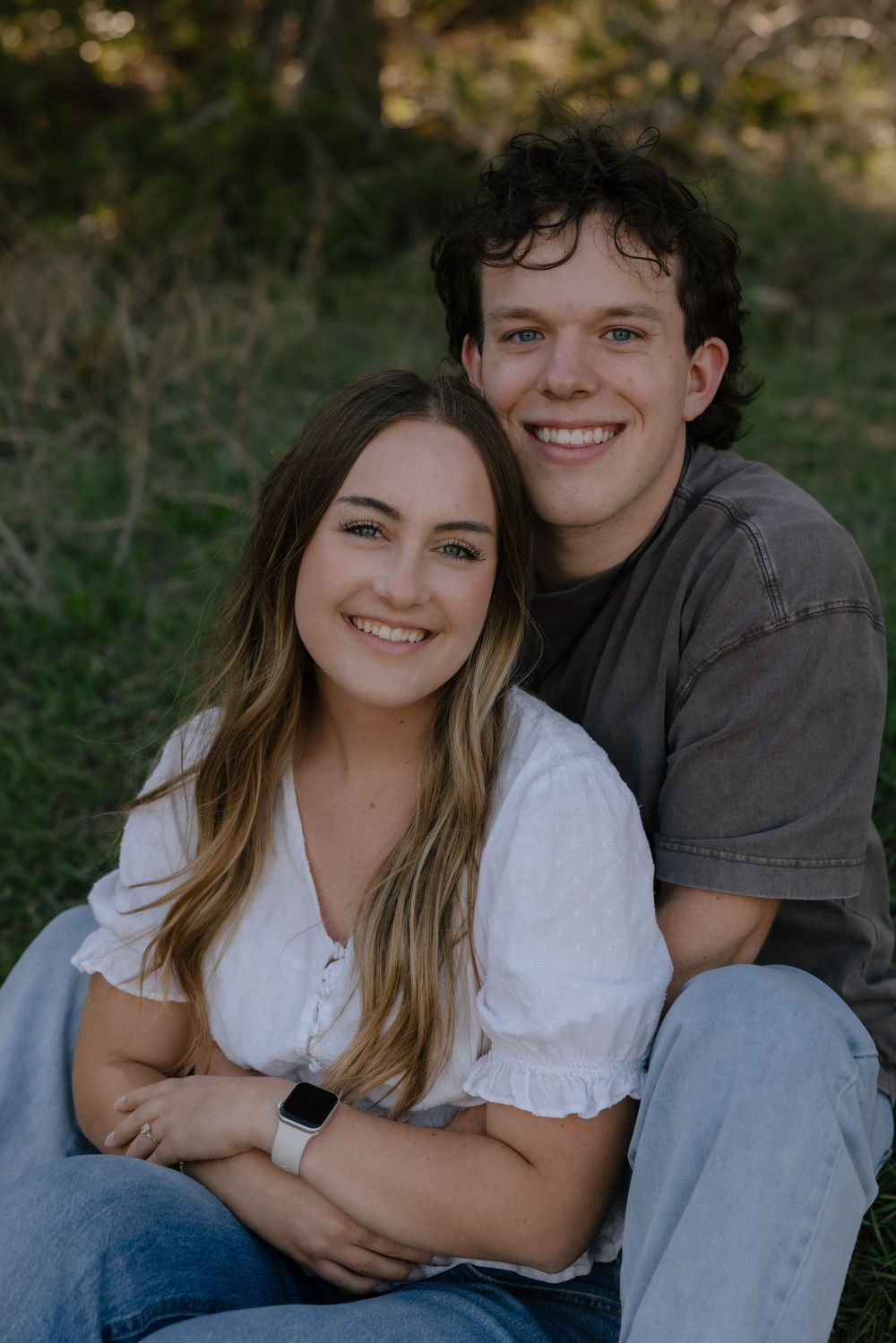 Smiling young couple sitting together in a green meadow, woman in white top and jeans with smartwatch.
