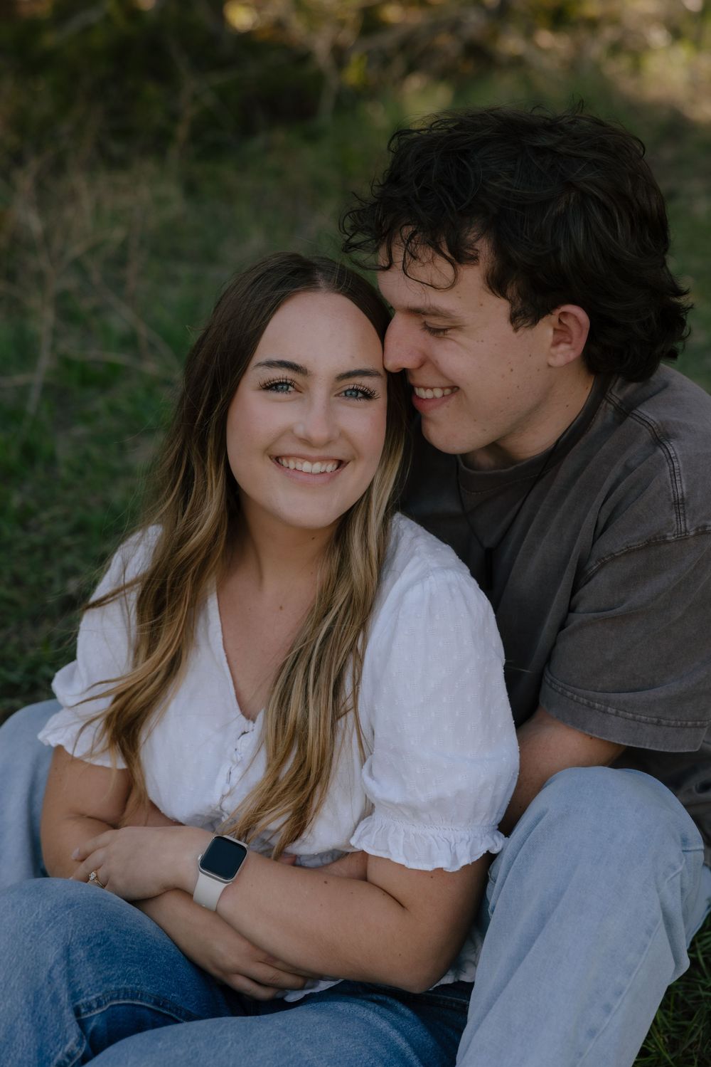 A smiling couple sits outdoors in nature, the woman in a white blouse and jeans leaning back against her partner.