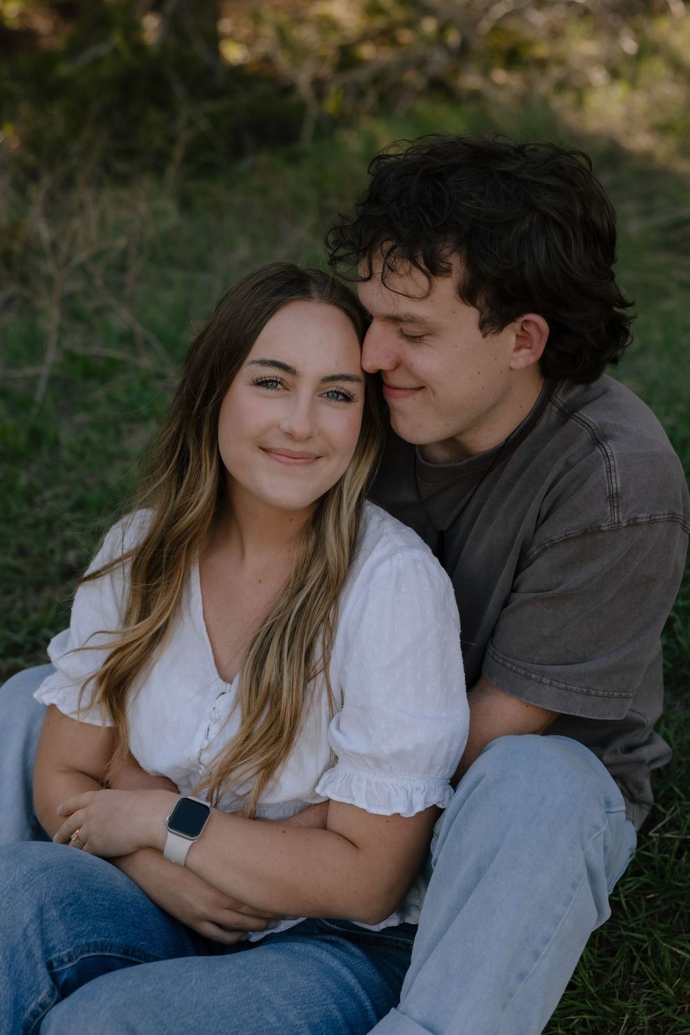 A smiling couple sits together in a grassy field, the man embracing the woman from behind affectionately.