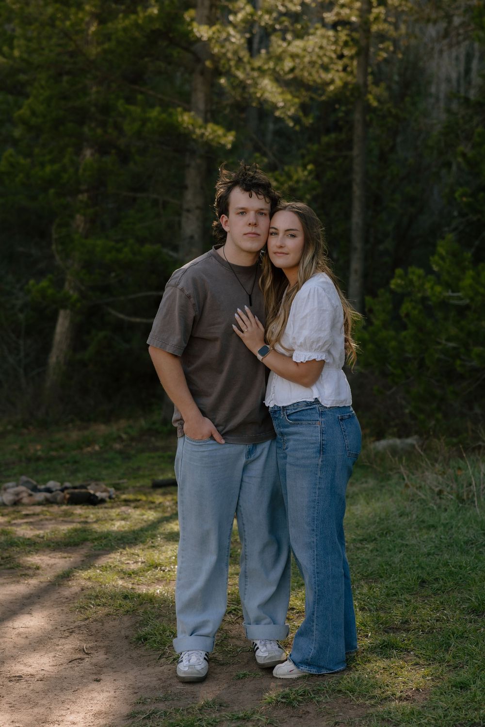 A couple embraces on a forest path, the woman in a white top and jeans leaning into the man in a brown t-shirt.