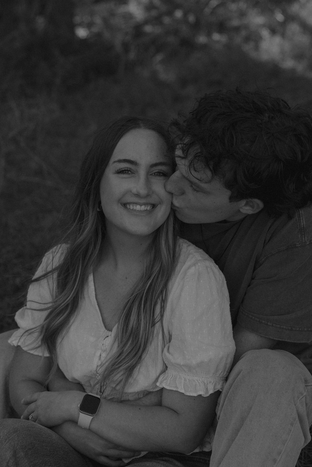Black and white photo of a smiling woman being kissed on the cheek by her partner while sitting outdoors.