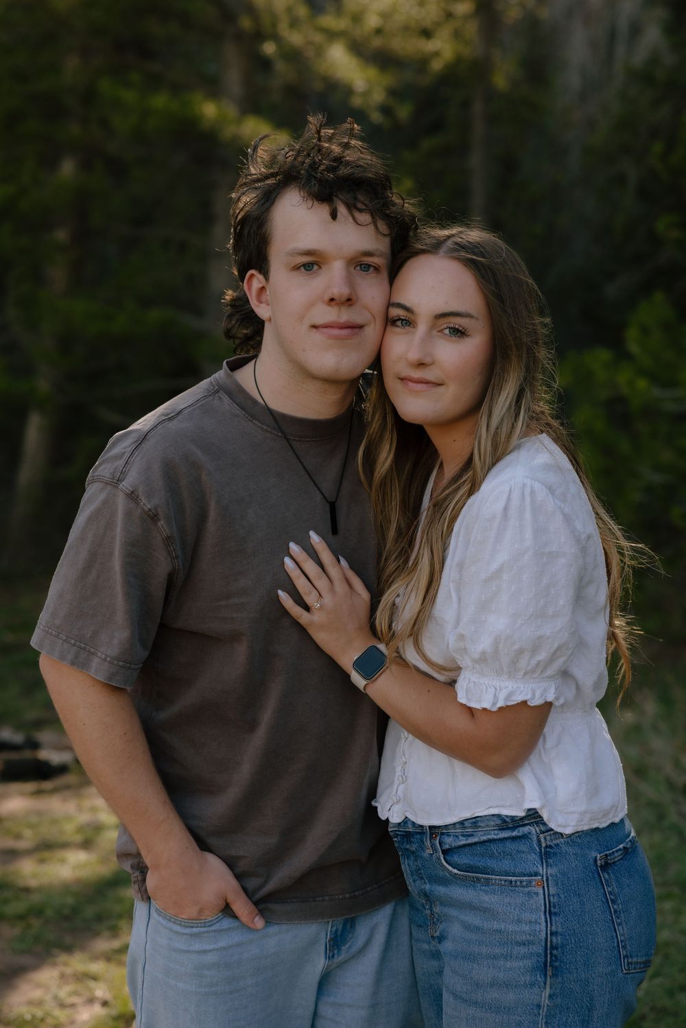 Young couple posing together in a forest, man in gray tee and woman in white blouse with jeans.